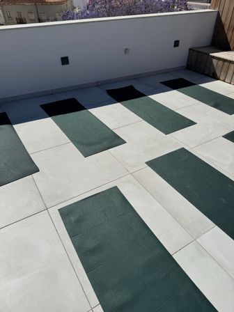 Sunlit rooftop terrace set up for rooftop yoga with evenly spaced dark green mats on pale tile, white low wall, wooden bench and purple flowering trees beyond.