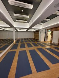 Empty indoor yoga studio with neat rows of navy mats on light wood floor, full-length mirror along one wall, recessed ceiling lighting and rustic wood accent wall