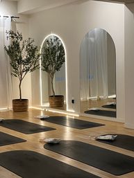 Minimal yoga/wellness studio interior with black mats and white eye pillows on light wood floor, arched LED-backlit mirrors, potted olive trees and sheer curtains