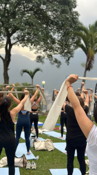 Group outdoor yoga/fitness class on mats in a grassy park, participants lifting stretch bands overhead with palm trees and misty mountains in the background.