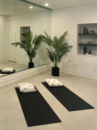Serene minimalist home yoga room with two black mats and white cushions on a polished concrete floor, large mirrored wall reflecting a potted palm and recessed shelves with decorative bottles.