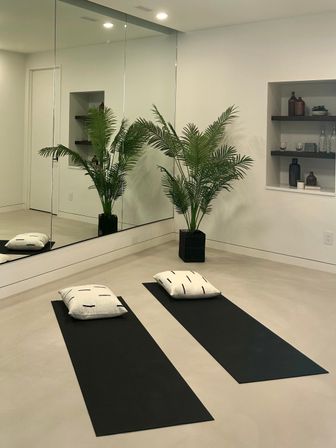 Serene minimalist home yoga room with two black mats and white cushions on a polished concrete floor, large mirrored wall reflecting a potted palm and recessed shelves with decorative bottles.