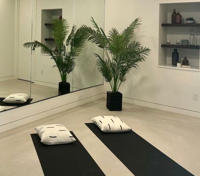 Serene minimalist home yoga room with two black mats and white cushions on a polished concrete floor, large mirrored wall reflecting a potted palm and recessed shelves with decorative bottles.
