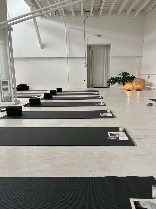 Minimalist white loft yoga studio with rows of black mats and blocks, candles and warm lanterns by a potted plant set for a calm yoga or meditation class