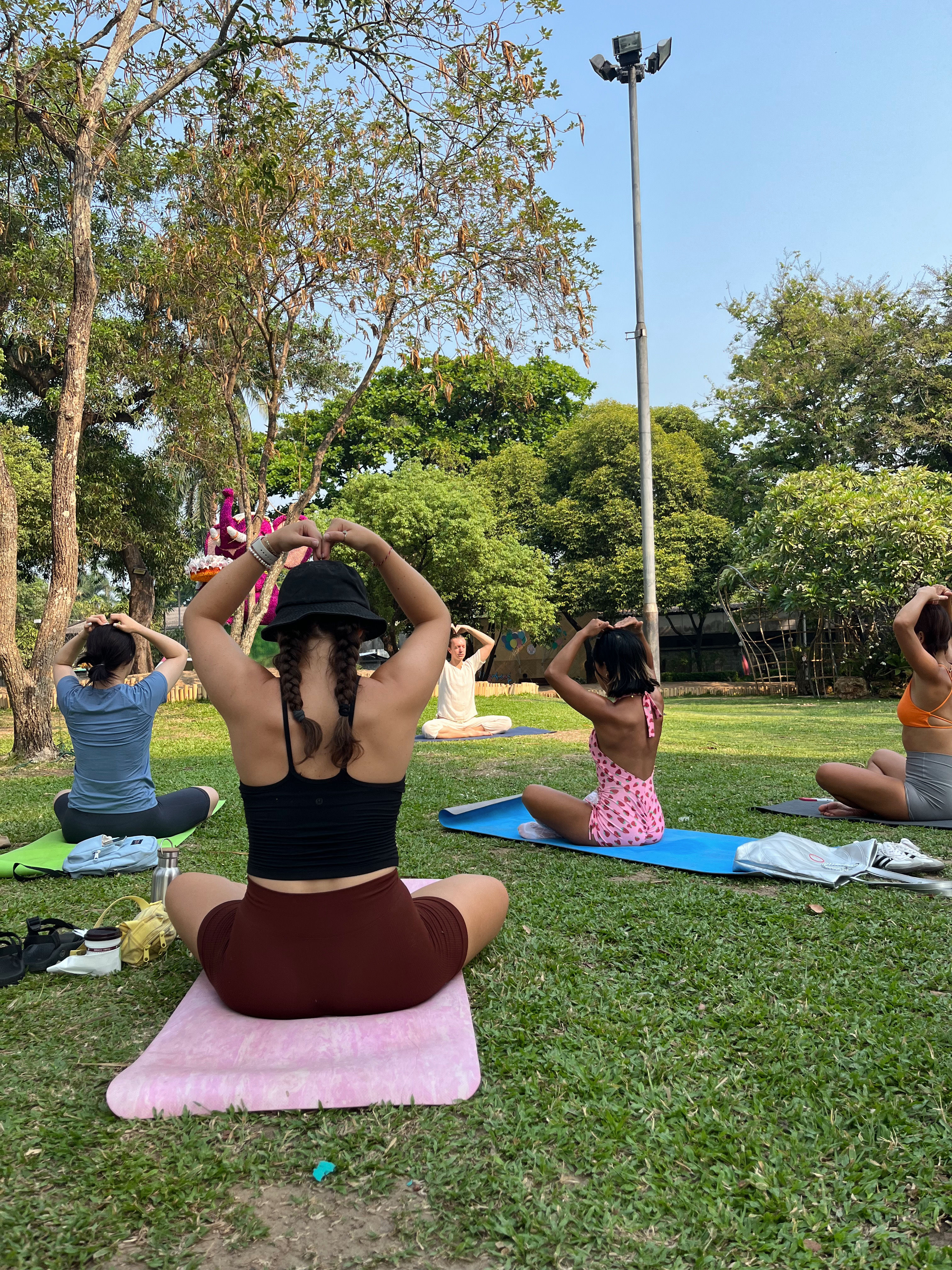 Sunny outdoor yoga class in a city park, participants seated on colorful mats on grass making heart shapes with their hands under trees and a tall light pole.