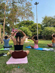 Sunny outdoor yoga class in a city park, participants seated on colorful mats on grass making heart shapes with their hands under trees and a tall light pole.