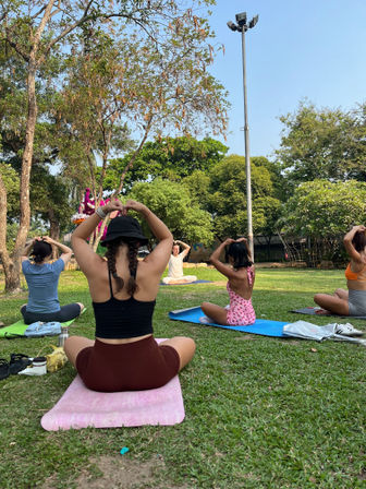 Sunny outdoor yoga class in a city park, participants seated on colorful mats on grass making heart shapes with their hands under trees and a tall light pole.