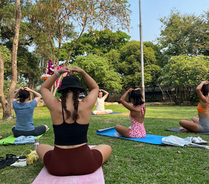 Sunny outdoor yoga class in a city park, participants seated on colorful mats on grass making heart shapes with their hands under trees and a tall light pole.