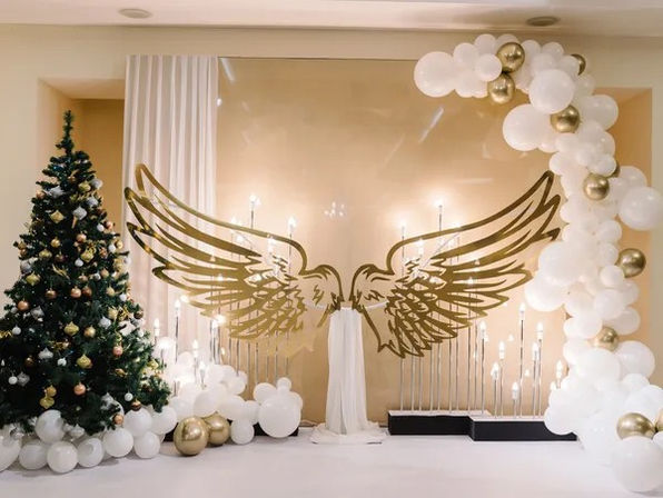 Festive indoor holiday photo backdrop with oversized gold angel wings, white-and-gold balloon arch, decorated Christmas tree, and glowing candles in an event space