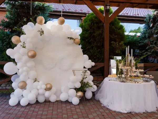 White and gold balloon arch framing a round white backdrop beside a tulle-covered dessert table with candles and sweets on a brick patio under a wooden pergola — outdoor wedding reception decor