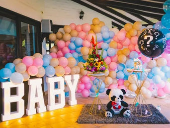 Indoor baby shower/gender-reveal setup with pastel pink, blue and gold balloon wall, illuminated "BABY" letters, floral centerpieces, panda plush and a black question‑mark reveal balloon.