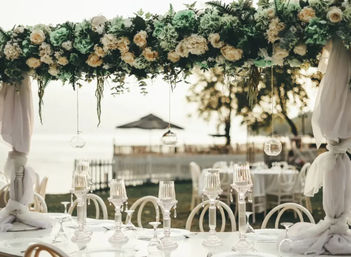 Elegant outdoor waterfront wedding table beneath a lush cream-and-green floral canopy, draped white fabric pillars, crystal candelabras and white place settings with a blurred beachside backdrop