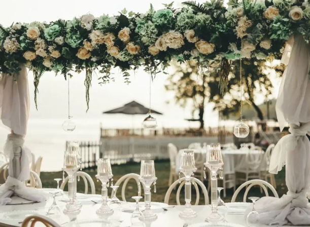 Elegant outdoor waterfront wedding table beneath a lush cream-and-green floral canopy, draped white fabric pillars, crystal candelabras and white place settings with a blurred beachside backdrop