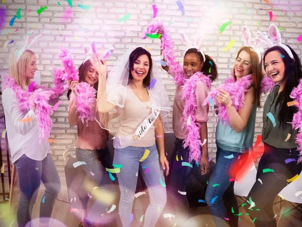 Bachelorette party: bride-to-be in veil and sash laughing with five friends wearing bunny ears and pink feather boas under colorful confetti in front of a brick wall.
