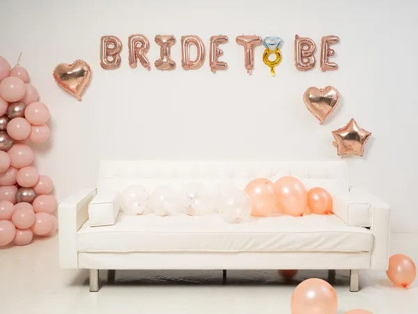 Rose-gold "BRIDE TO BE" foil balloons with heart and star accents above a white sofa surrounded by pink and peach balloons for a stylish bridal shower backdrop