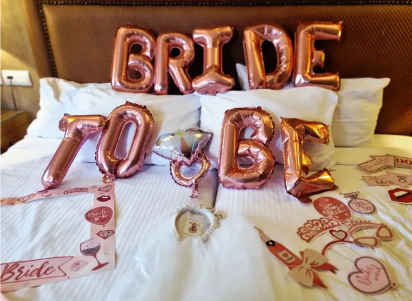 Rose-gold foil "BRIDE" and "TO BE" balloons on a hotel bed with tiara, bridal sash and bachelorette party props — festive bridal celebration setup.