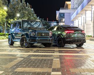 Two dark luxury SUVs — a customized Mercedes G-Class and a Lamborghini Urus — parked on a wet patterned downtown plaza at night with city lights and a modern office building in the background.