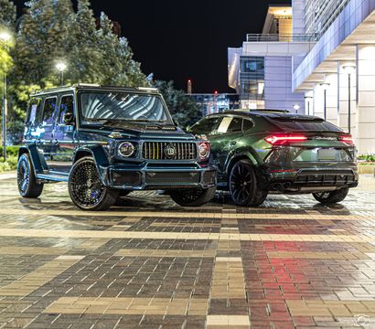 Two dark luxury SUVs — a customized Mercedes G-Class and a Lamborghini Urus — parked on a wet patterned downtown plaza at night with city lights and a modern office building in the background.