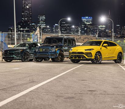 Three luxury SUVs parked in an urban night parking lot with a glowing downtown skyline, a bright yellow sports SUV in front.