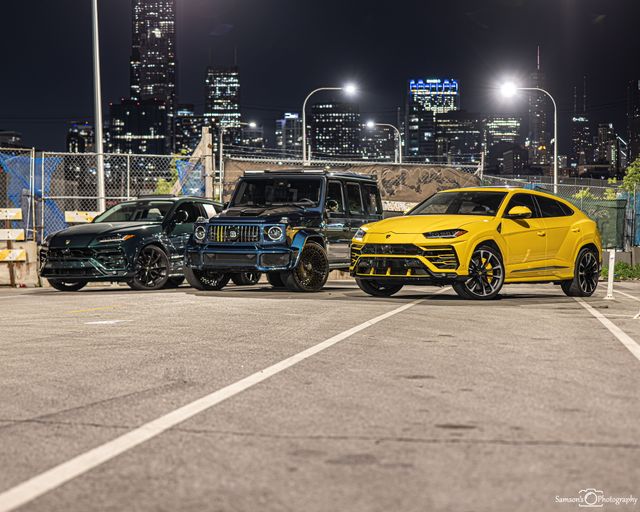 Three luxury SUVs parked in an urban night parking lot with a glowing downtown skyline, a bright yellow sports SUV in front.