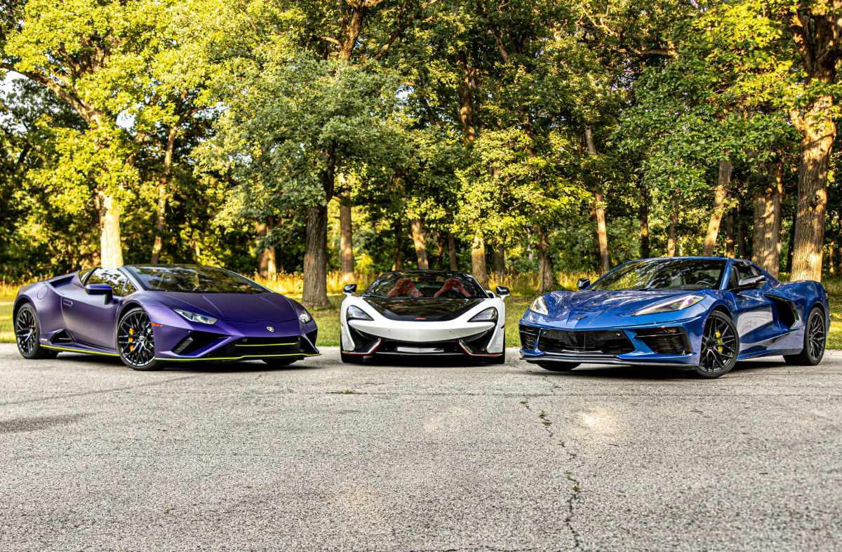 Three exotic sports cars — purple, white, and blue — parked on a sunlit paved clearing surrounded by leafy trees in a park-like setting.