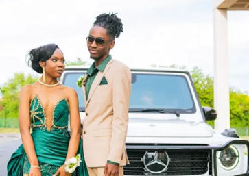 Glamorous prom couple posing by a white luxury SUV in a parking lot — woman in an emerald green beaded gown with corsage and pearl necklace, man in a tan suit with green shirt and sunglasses, stylish outdoor portrait.