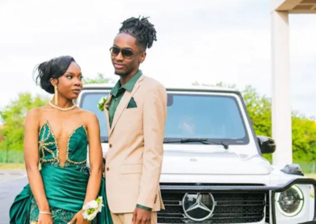 Glamorous prom couple posing by a white luxury SUV in a parking lot — woman in an emerald green beaded gown with corsage and pearl necklace, man in a tan suit with green shirt and sunglasses, stylish outdoor portrait.