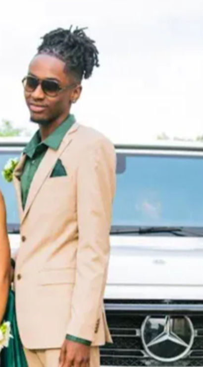 Glamorous prom couple posing by a white luxury SUV in a parking lot — woman in an emerald green beaded gown with corsage and pearl necklace, man in a tan suit with green shirt and sunglasses, stylish outdoor portrait.