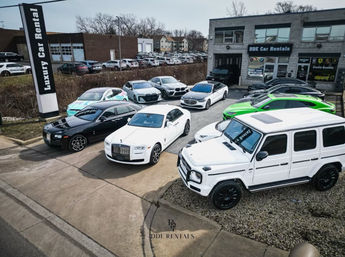 Aerial view of a suburban luxury car rental lot featuring a white Mercedes G‑Wagon, white Rolls‑Royce, silver sedans and a neon green sports car parked outside a garage.
