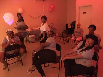 Group of women at an indoor bachelorette party in pink lighting, seated on folding chairs with balloons, snacks table and a "miss to mrs" banner