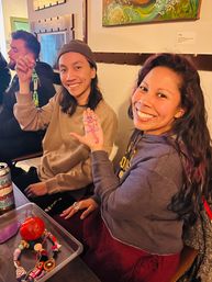 Two smiling friends at a cozy indoor craft table proudly showing colorful handmade beaded keychains and charms, with a tray of craft supplies and a canned drink visible
