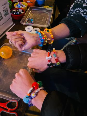 Close-up of several wrists wearing colorful handmade beaded bracelets with charms and pom-poms on a craft table next to bowls of beads and string
