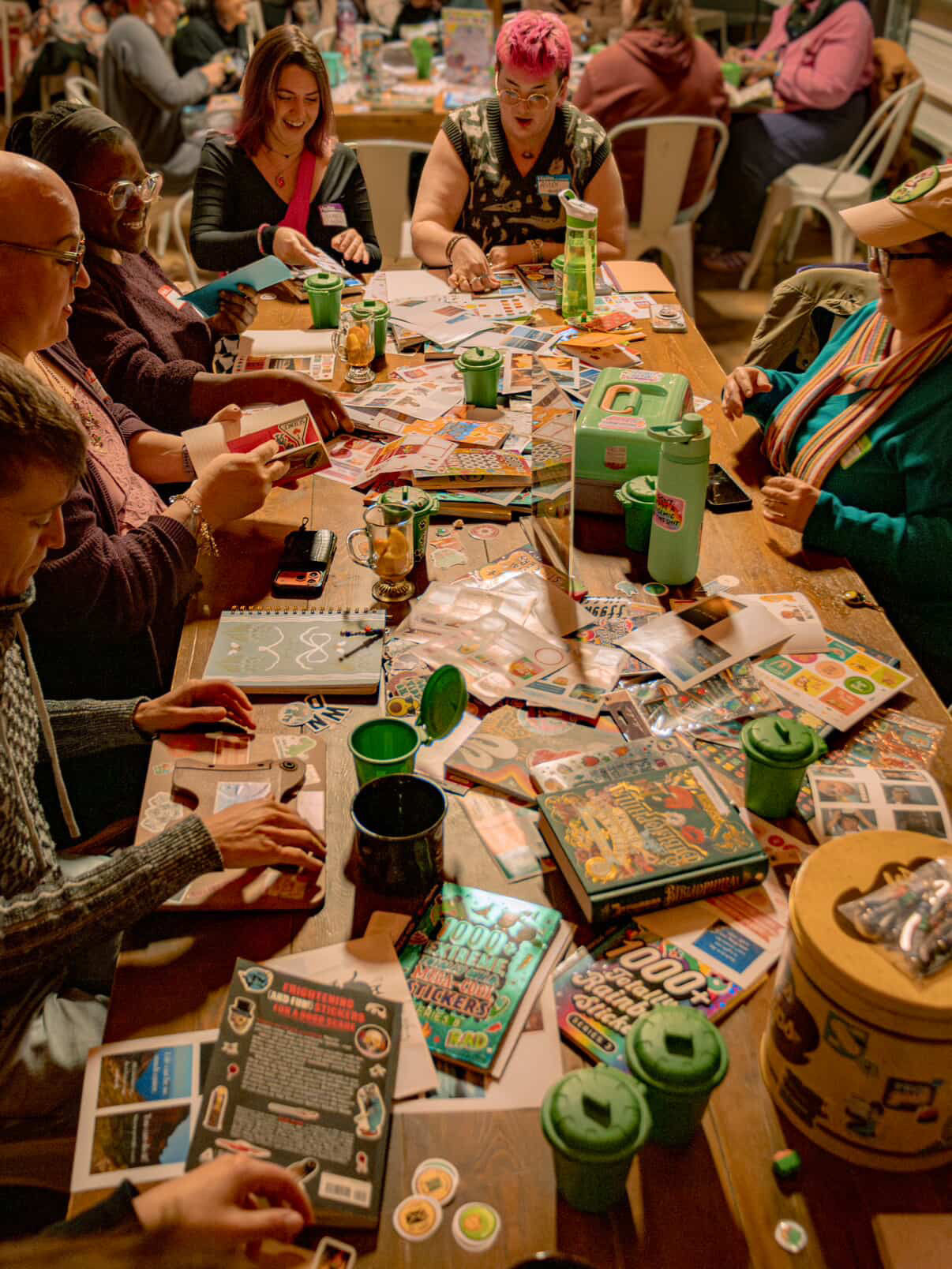 Community craft night with people gathered around a long wooden table making sticker collages and paper projects, books, scissors, stickers and green drink tumblers scattered across the workspace.
