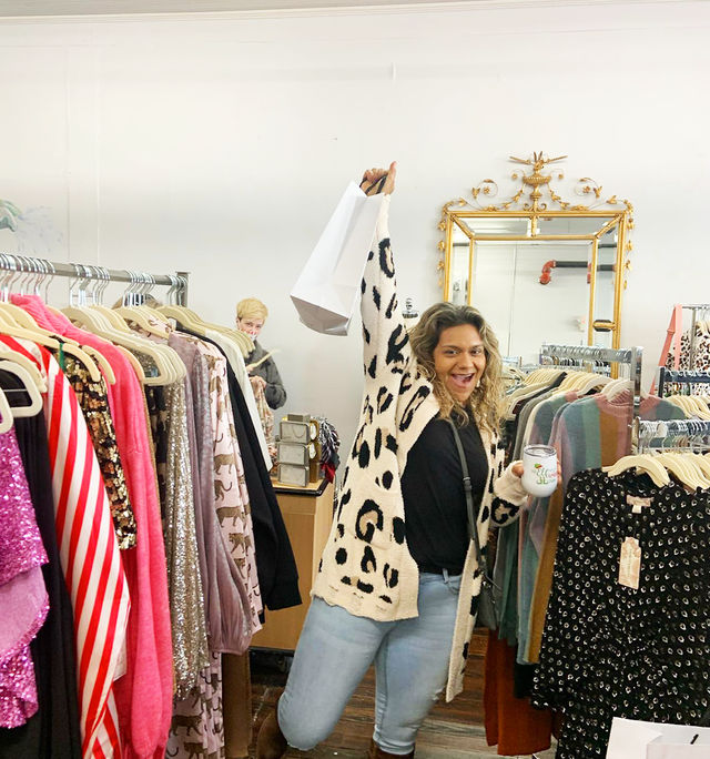 Cheerful woman in a leopard-print cardigan holding a shopping bag and drink inside a bright women’s clothing boutique, posing among colorful clothing racks and a gold-framed mirror.