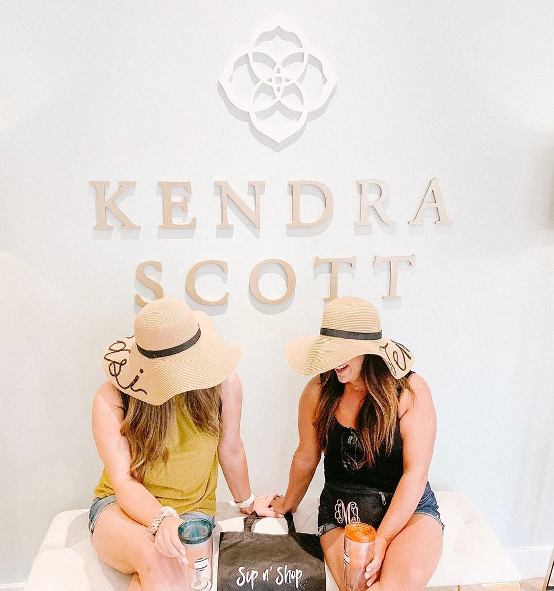 Two women in floppy sun hats sit on a bench in a bright jewelry boutique, laughing and holding tumblers with a 'Sip n' Shop' bag in front of a decorative logo wall.