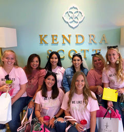 Eight smiling women in matching pink 'We Like to Party' shirts pose inside a pastel mint jewelry boutique with gold lettering and floral logo, holding shopping bags and colorful tumblers — cheerful shopping celebration.