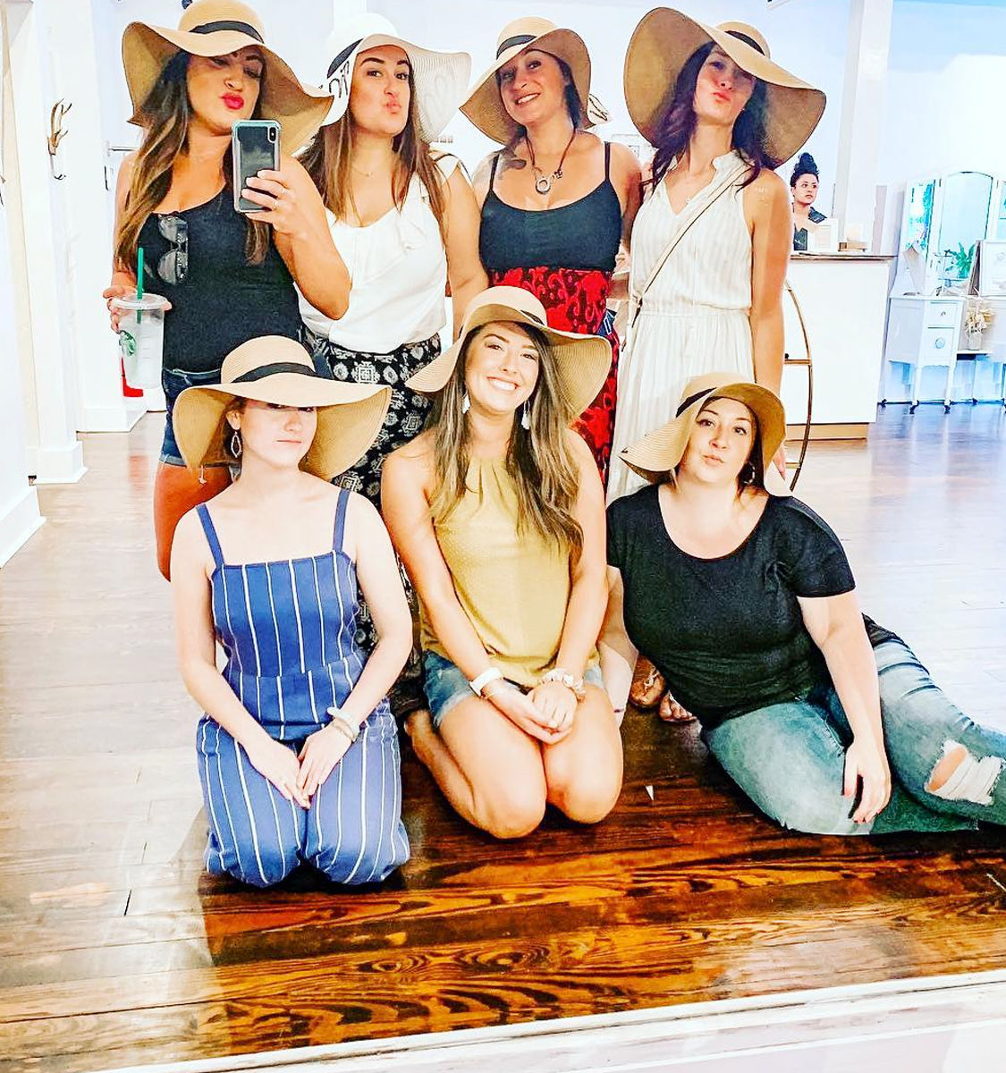 Seven women wearing floppy sun hats pose for a mirror selfie inside a bright boutique with hardwood floors — playful hat-shopping moment.