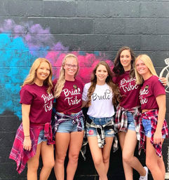 Five friends pose smiling in front of a colorful spray-painted brick mural; bride in a white “Bride” tee flanked by four bridesmaids in matching maroon “Bride Tribe” shirts, denim shorts and flannel shirts tied at their waists — playful bachelorette celebration shot.