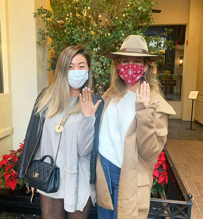 Two masked women in an indoor holiday courtyard — one in a red snowflake mask and hat, the other in a white mask and leather jacket — both showing rings on their left hands in front of a lit potted tree and poinsettias.