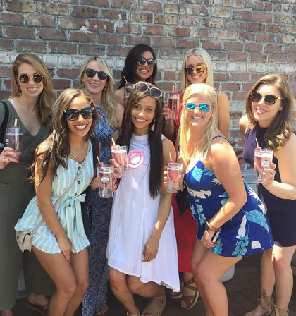 Eight women in summer dresses and sunglasses smiling and holding pink cocktails with popsicle garnishes, posing outdoors against a brick wall.