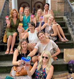Cheerful group of women on brownstone townhouse steps in summer outfits, toasting with colorful travel tumblers and surrounded by shopping bags on a city stoop.