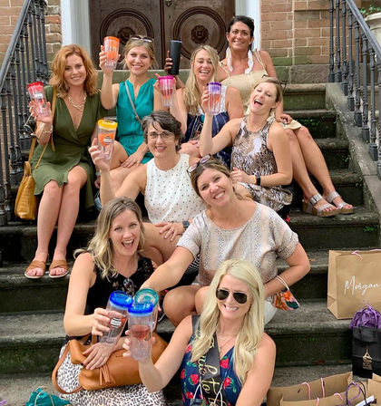 Cheerful group of women on brownstone townhouse steps in summer outfits, toasting with colorful travel tumblers and surrounded by shopping bags on a city stoop.