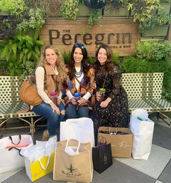 Three friends smiling on a woven bench in front of a vertical garden and wooden sign, holding colorful drinks with multiple shopping bags and gift totes at their feet on an outdoor patio.