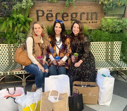 Three friends smiling on a woven bench in front of a vertical garden and wooden sign, holding colorful drinks with multiple shopping bags and gift totes at their feet on an outdoor patio.
