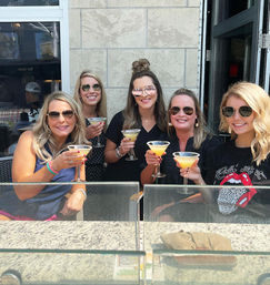 Five women friends wearing sunglasses smile and toast with yellow cocktails in martini glasses on a sunny outdoor patio bar against a stone wall backdrop.