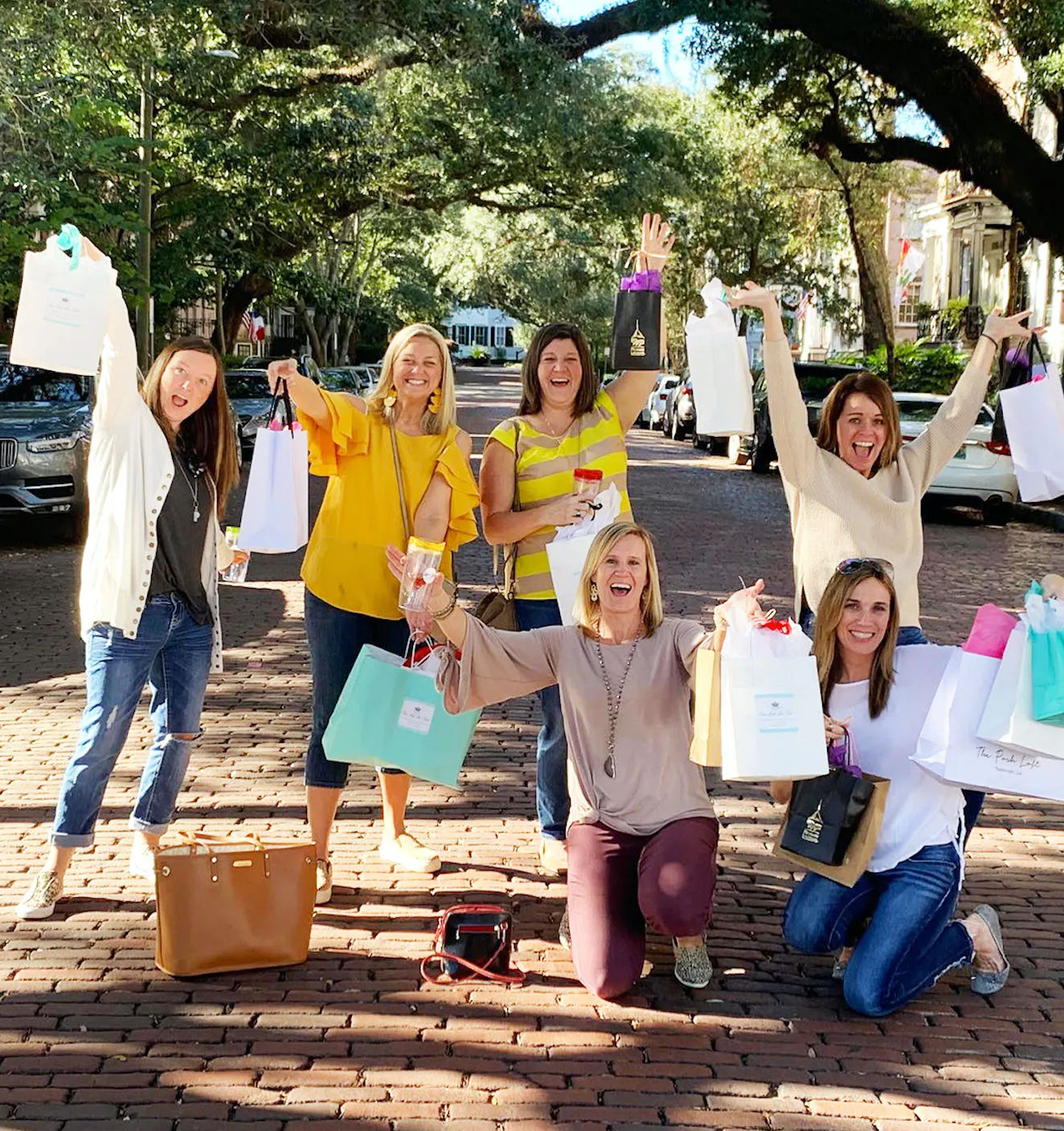 Six women on a tree-lined brick street celebrating a girls' day out, smiling and holding colorful shopping bags under a sunny canopy of mature trees.
