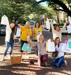 Six women on a tree-lined brick street celebrating a girls' day out, smiling and holding colorful shopping bags under a sunny canopy of mature trees.