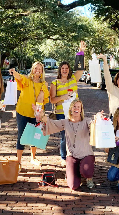 Six women on a tree-lined brick street celebrating a girls' day out, smiling and holding colorful shopping bags under a sunny canopy of mature trees.