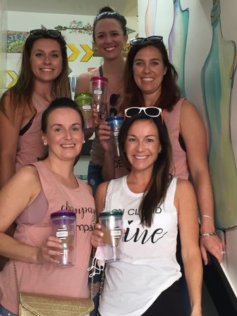 Five smiling women holding colorful wine tumblers during a girls' day out in a bright indoor space with a painted mural