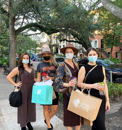 Four masked women in dresses and hats posing on a tree-lined historic downtown sidewalk, holding shopping bags and tumbler drinks beneath a moss‑draped oak canopy and brick townhouses.